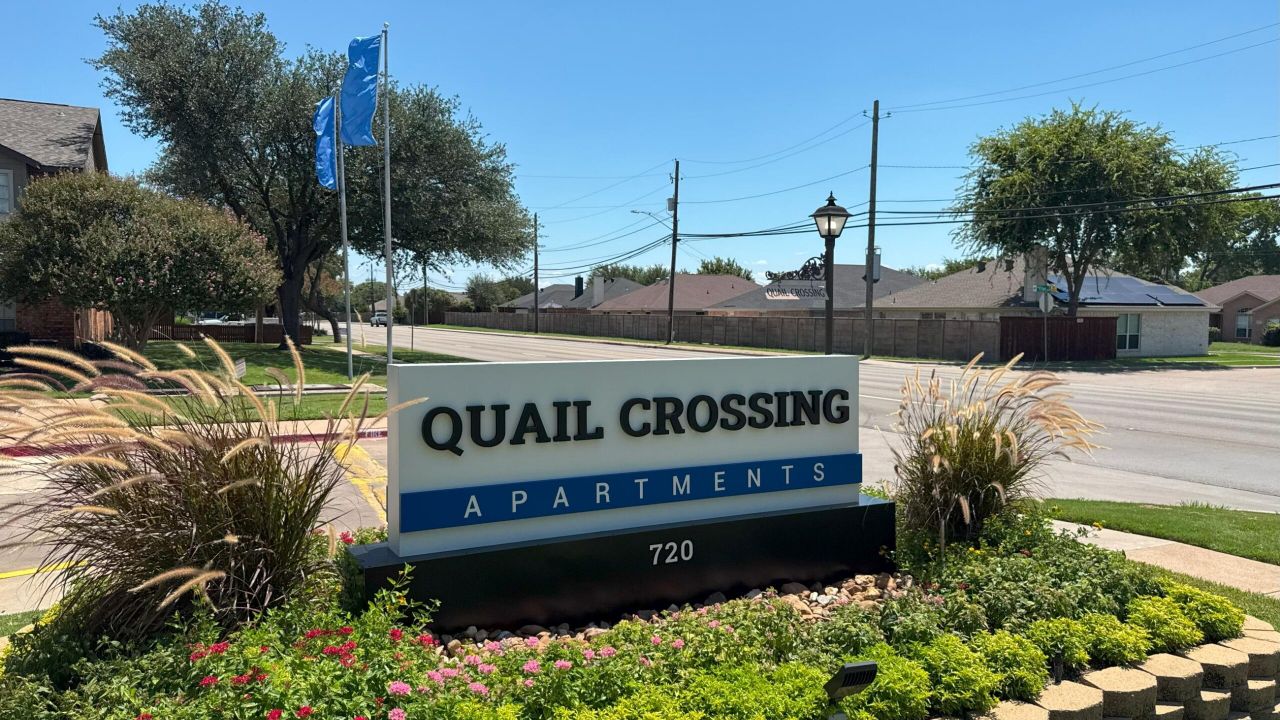 Sign reading "Quail Crossing Apartments" with flowers and trees in the background.