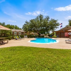 Quail Crossing Apartments Sunny outdoor pool area with loungers, picnic tables, and umbrellas surrounded by trees and a grassy lawn.