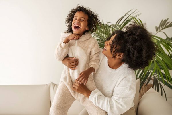Quail Crossing A child in a fluffy outfit laughs while being held by a smiling adult; they are in a bright room with plants.