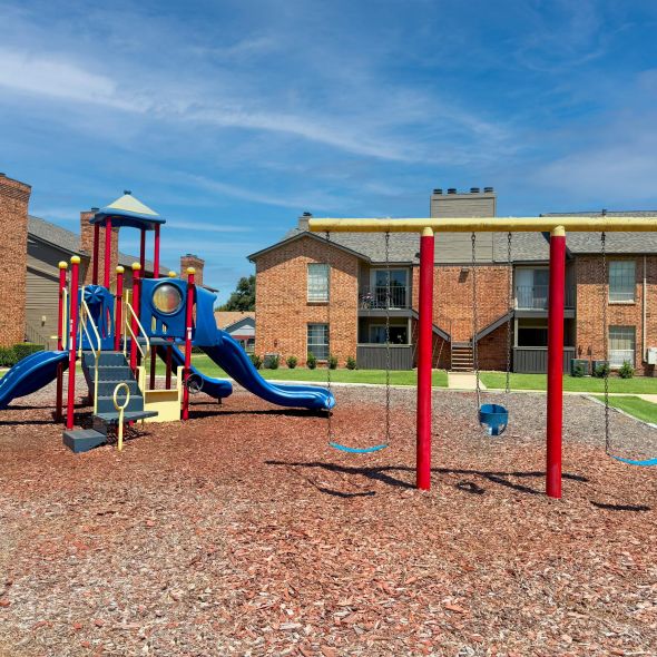 Quail Crossing Playground with slides and swings in front of brick apartment buildings under a clear blue sky.